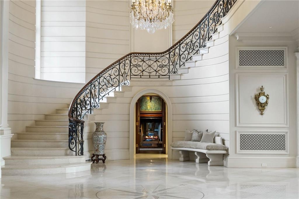 Marble entry hall of LONGVIEW with a sweeping iron-railed staircase, a crystal chandelier, and a tall arched doorway leading to a fireplace beyond