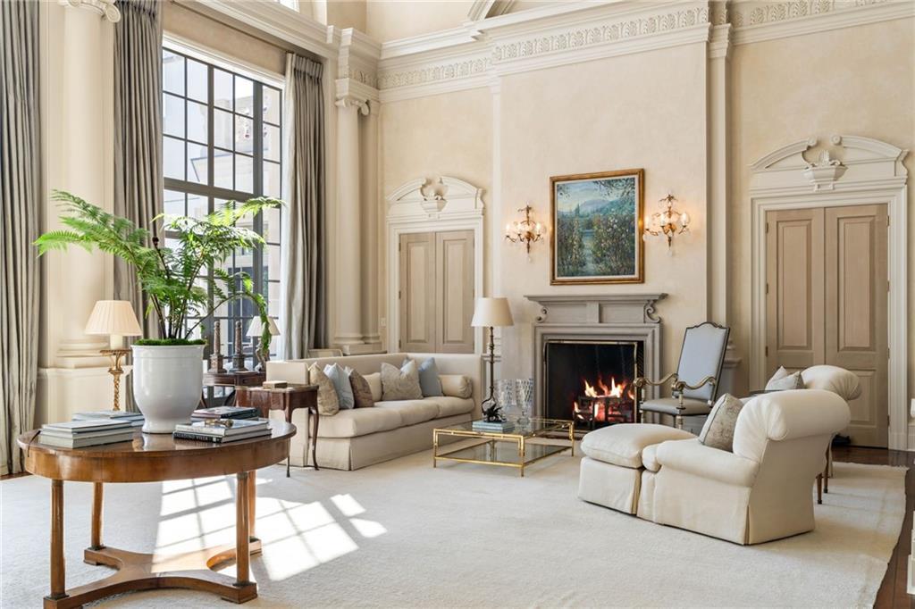 Formal two-story living room at LONGVIEW with Venetian plaster walls, a lit fireplace, cream upholstery with blue accent pillows, and tall mullioned windows letting in afternoon light