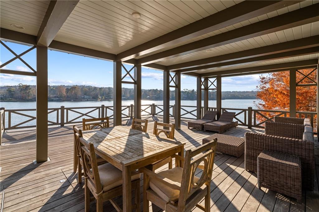 Covered upper deck of the LONGVIEW boathouse looking across Lake Oliver, with wood plank floors, an X-brace railing, a teak dining table for six, and an orange-leafed tree at the far end