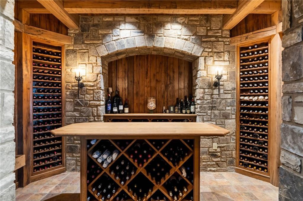 Stone-walled wine cellar at LONGVIEW with a heavy timber-beamed ceiling, two tall bottle racks under sconces, and a wood tasting table set with bottles and decanters